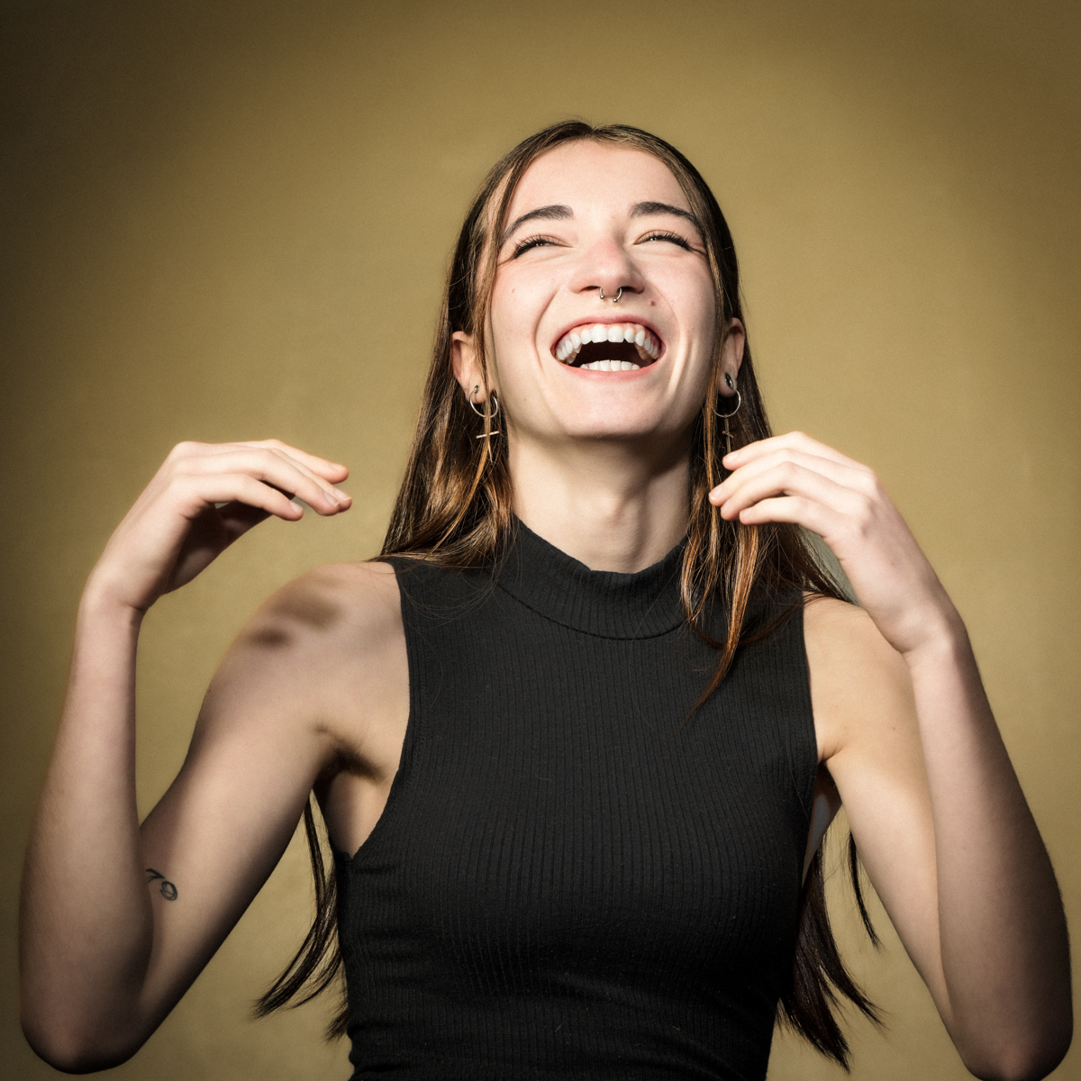 Jeune femme aux longs cheveux, débardeur noir, boucles d'oreilles croix, fond beige.