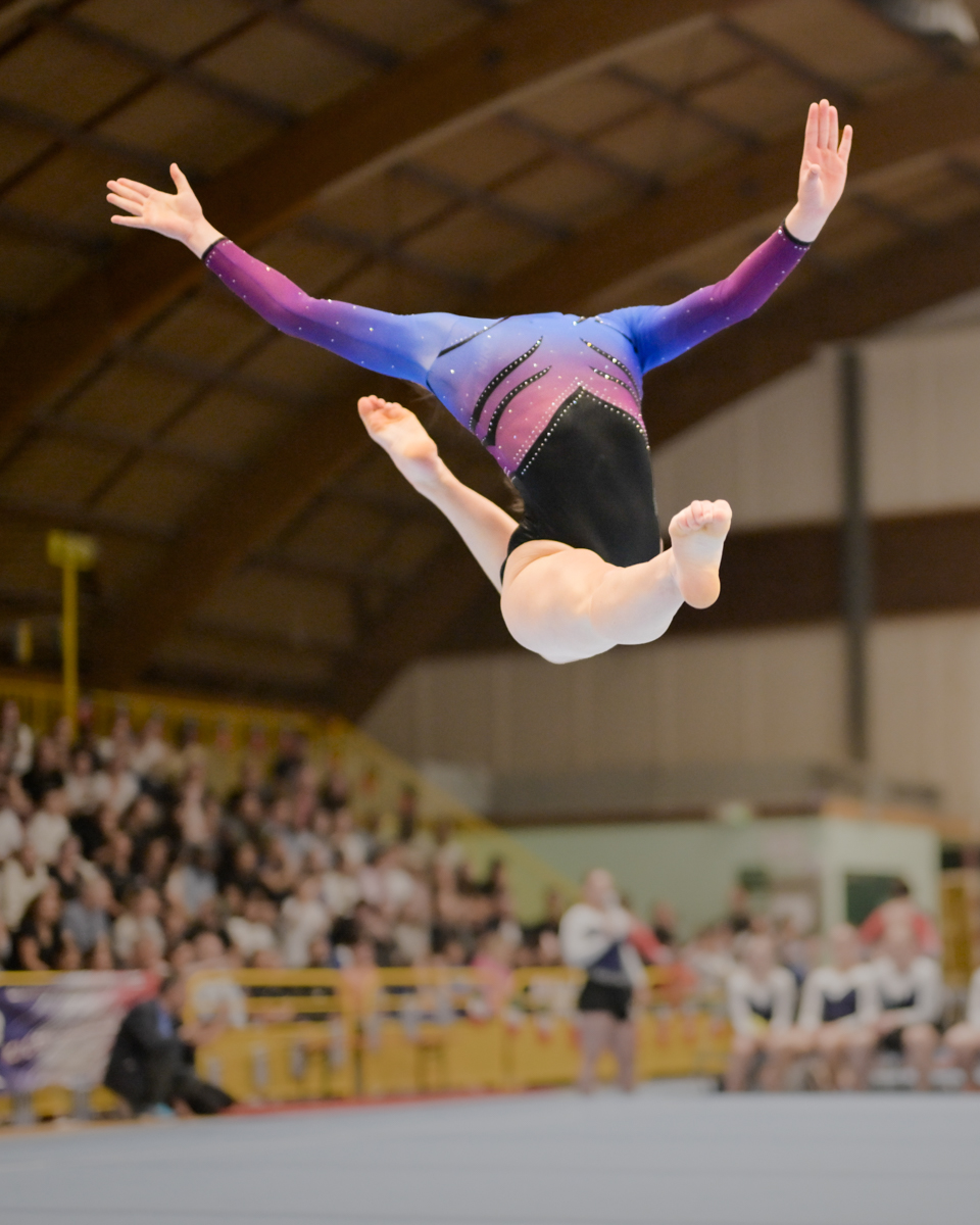 Gymnaste en tenue colorée effectuant un saut acrobatique en salle devant un public.