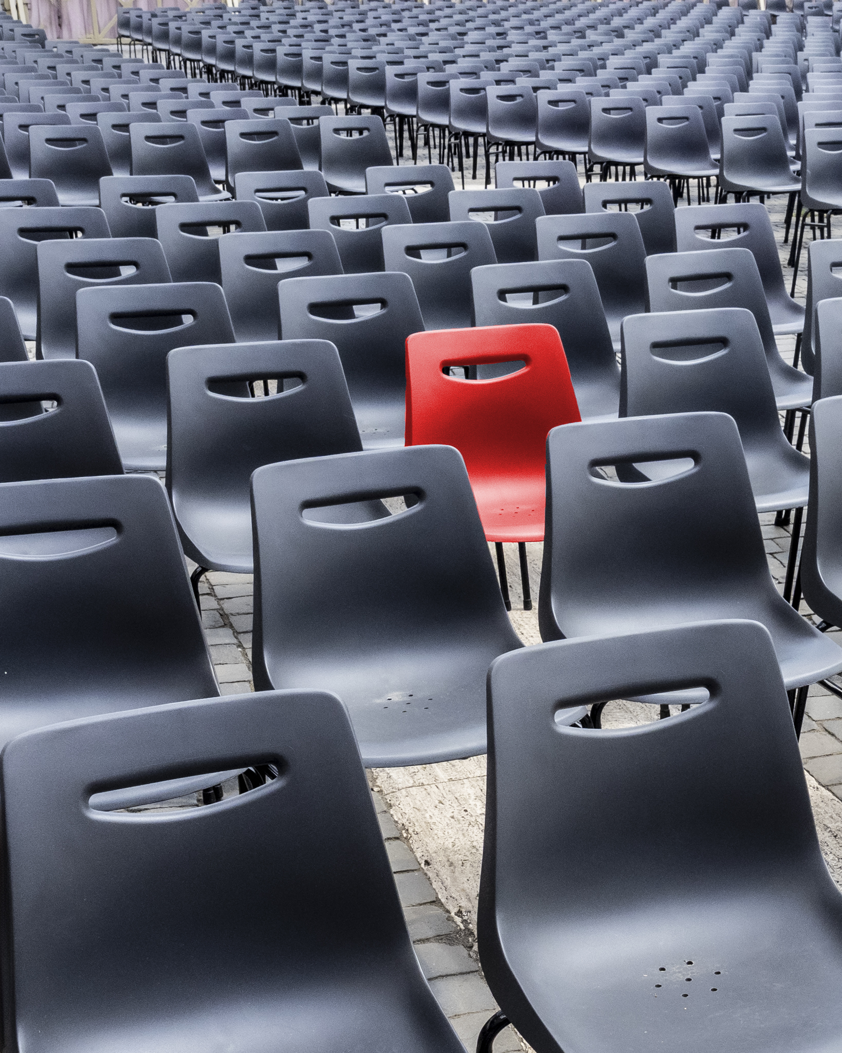 Rangée de chaises en plastique noir avec une seule chaise rouge au centre sur sol pavé.