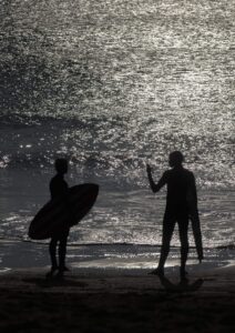 Surfeurs en silhouette avec reflets sur l’eau. Deux surfeurs en silhouette sur une plage, chacun avec sa planche; eau scintillante.