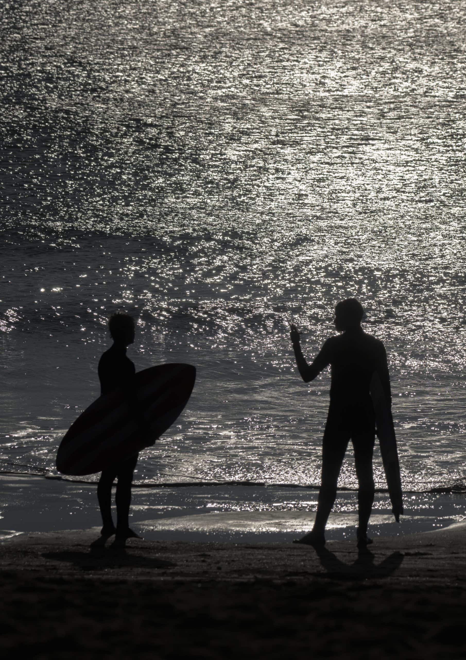 Deux surfeurs en silhouette sur une plage, chacun avec sa planche; eau scintillante.