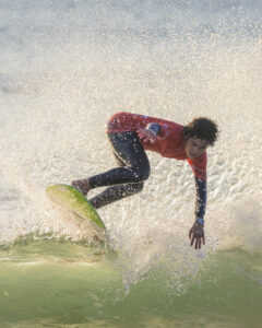 Surfeur en combinaison noire sur planche verte Surfeur en combinaison noire et haut rouge sur planche verte avec éclaboussures d'eau.
