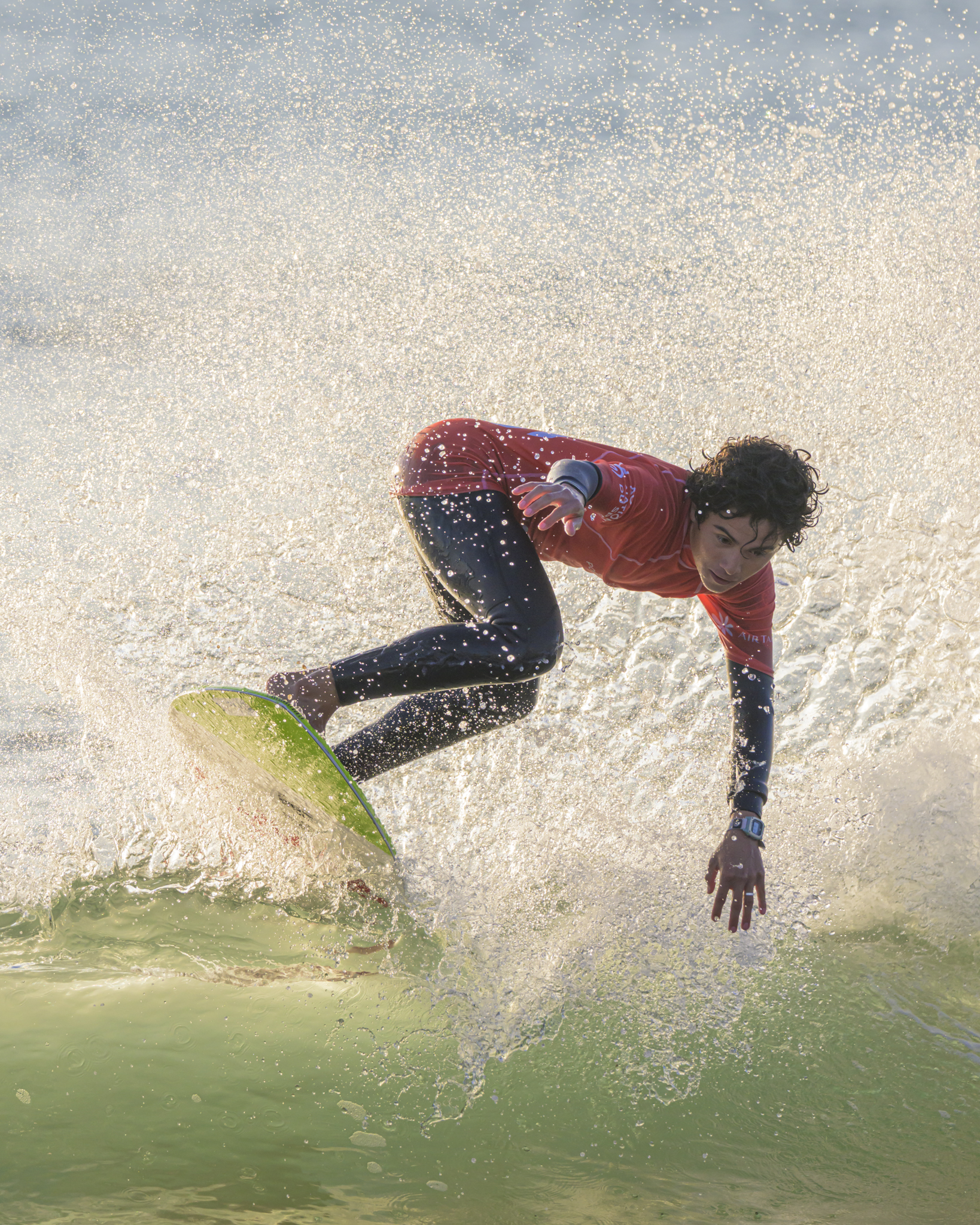 Surfeur en combinaison noire et haut rouge sur planche verte avec éclaboussures d'eau.