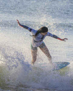 Surfeuse en tenue à manches longues équilibre sur planche bleue dans éclaboussures d'eau.