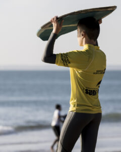 Jeune surfeur en combinaison et maillot jaune tenant une planche face à la mer sur la plage.