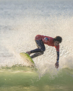 Surfeur en combinaison noire et haut rouge réalisant une manœuvre sur une vague verte.
